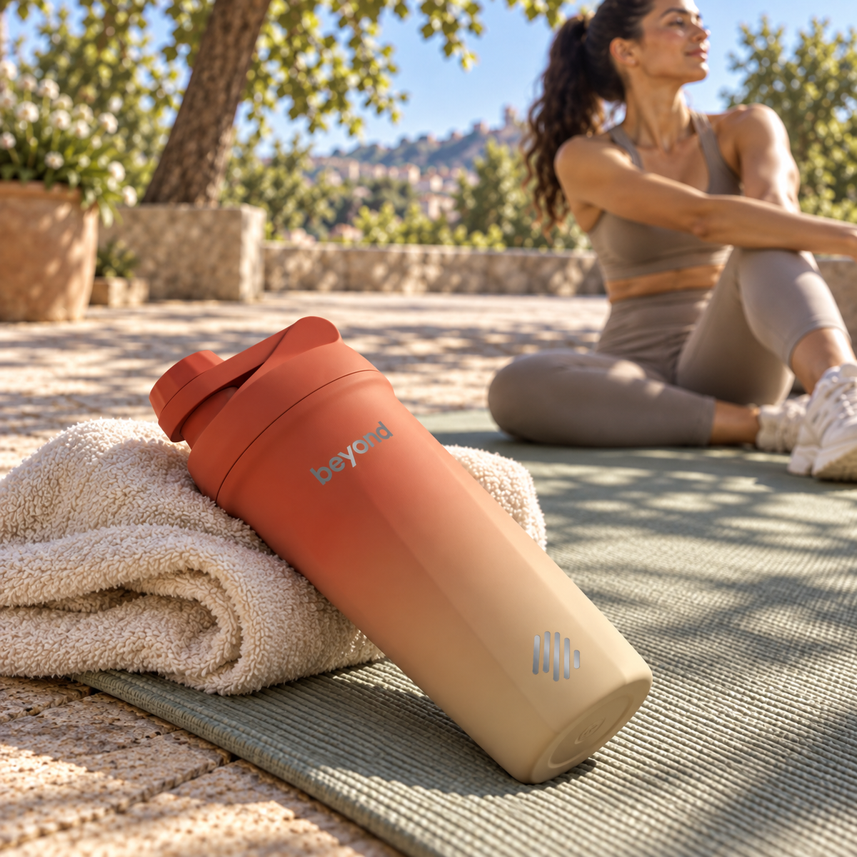 A Beyond brand shaker bottle with a terracotta-to-beige gradient, resting on a white towel on an outdoor yoga mat with a woman in athletic wear blurred in the background.