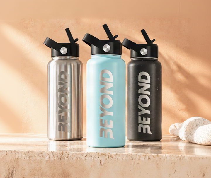 Three insulated stainless steel water bottles with black flip-top lids stand on a stone surface against a warm beige wall with soft shadows. The bottles are silver, light blue, and dark gray/black, each printed with the word 'BEYOND' vertically.